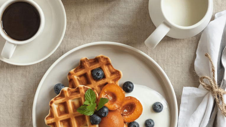 Overhead view of waffles topped with poached apricots, blueberries, yogurt, and mint, served with black coffee, milk, and a bowl of blueberries.