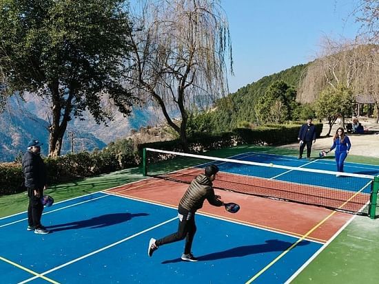 Guests playing pickle ball on vibrant blue and red court surrounded by mountain scenery and lush gardens at The Manor Wellness & Sports Hotel