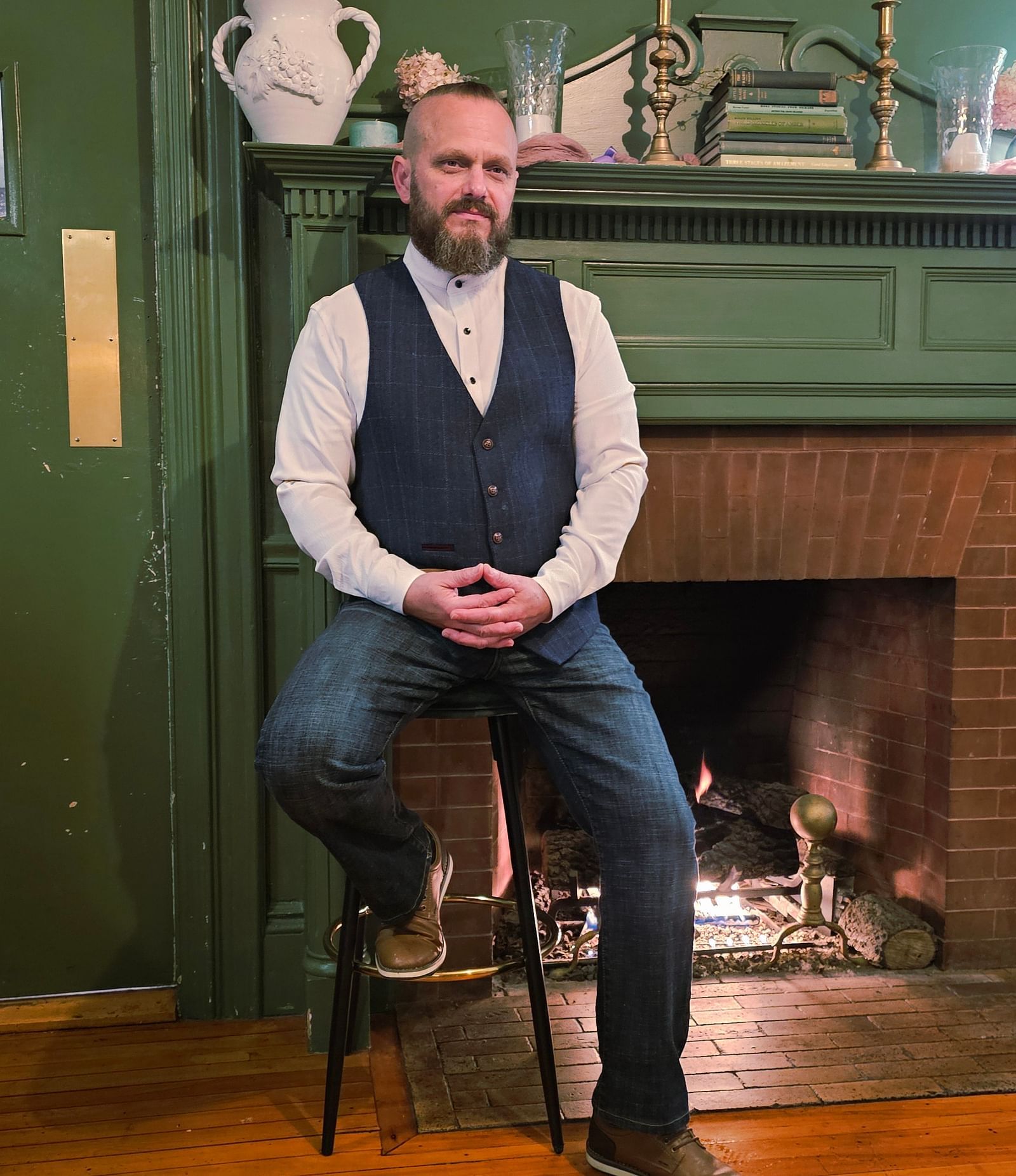 Chef Tom E. White seated on a stool in front of a green fireplace, wearing a dark vest, white shirt, and jeans.