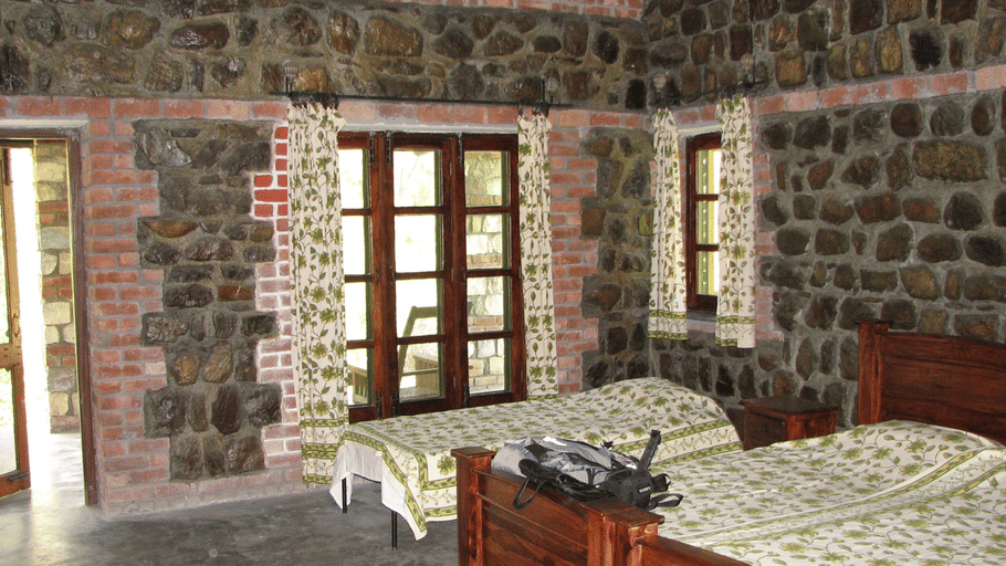 A spacious rustic cabin bedroom featuring Exposed stone and brick walls, wooden twin beds, and large windows with patterned curtains.