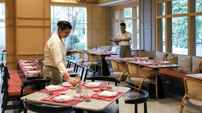 A restaurant interior at Heritage Village Resorts & Spa, Manesar, with staff arranging table settings, placemats, and folded napkins, with another staff member standing near the windows.