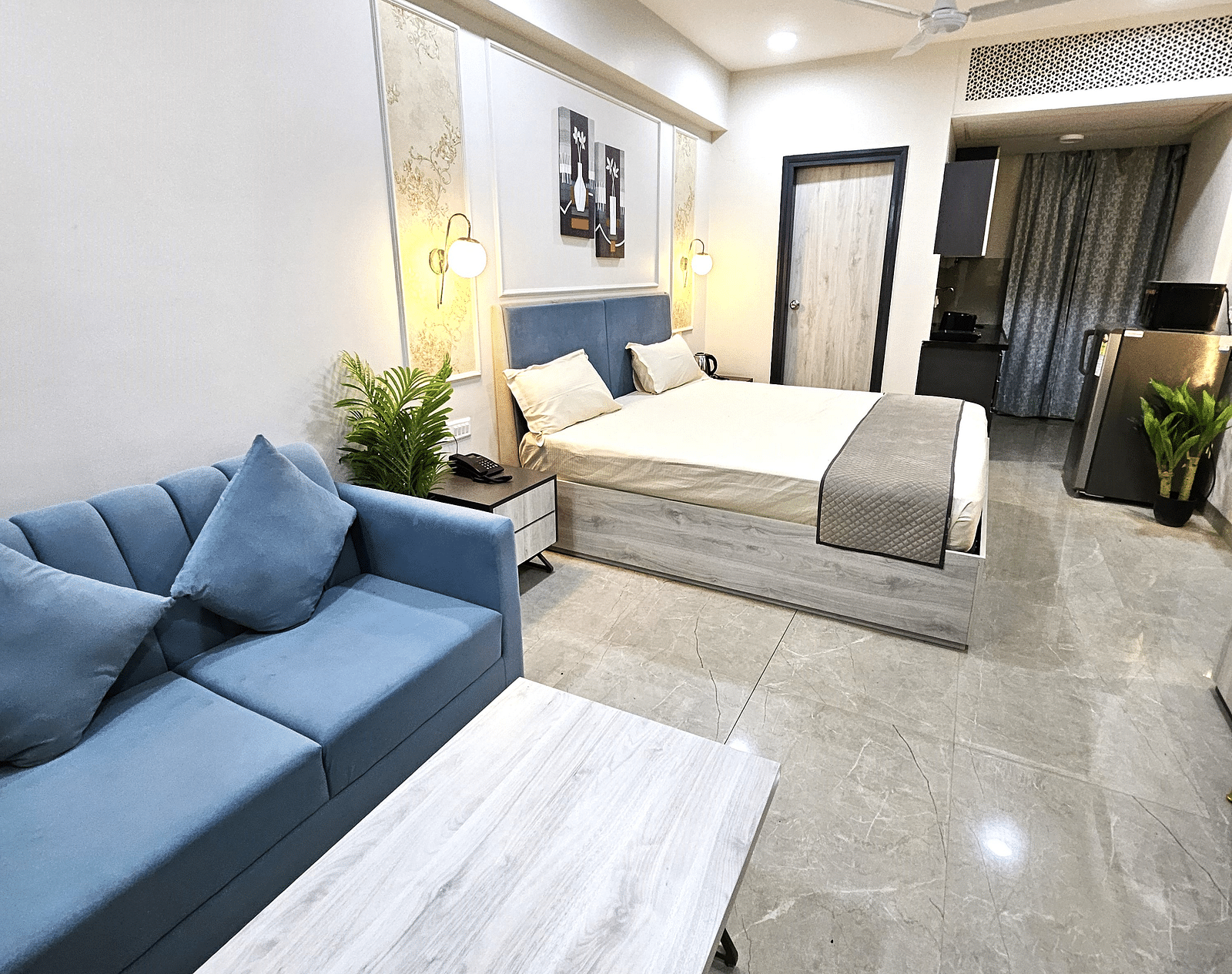 Modern studio room featuring a bed, blue sofa with matching cushions, wooden coffee table, kitchenette with appliances, and warm lighting accents - Urban Suites by BluSalzz, Jaipur