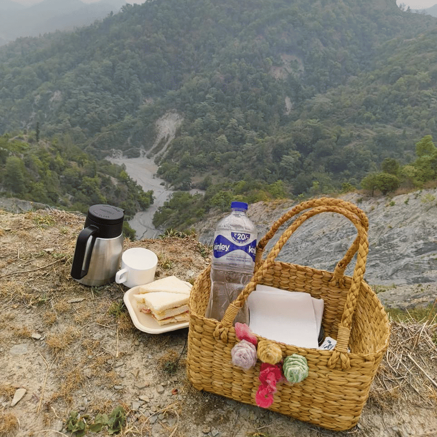 A picnic basket, flask, 1 bottle and snacks resting on a hill overlooking lush mountains.