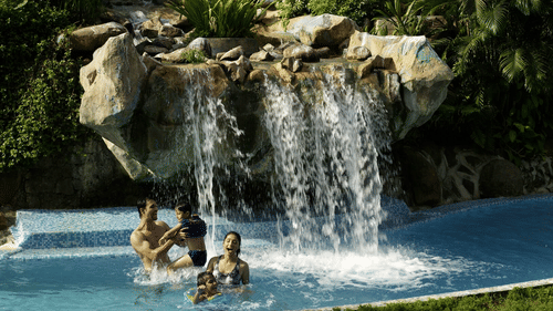 A waterfall flowing over rocks into a swimming pool with surrounding tropical vegetation and people in the pool at he Retreat Hotel and Convention Centre, one of the best hotels in Mumbai.