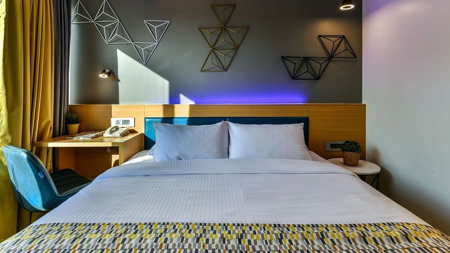 A view from the foot of a spacious bed, featuring a bedside table on one side and a work desk with a chair on the other, illuminated by cool lighting at Indie Stays, BKC Kalina.