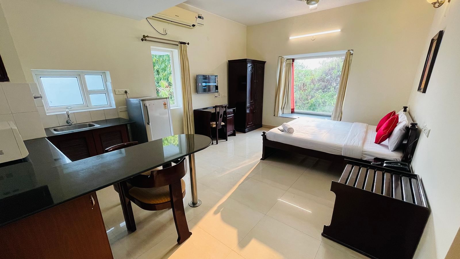 A studio room featuring a double bed with red accents, a desk, a chair, a kitchenette with dining table, chair, and a refrigerator at Sanctum Suites Indiranagar, Bangalore.