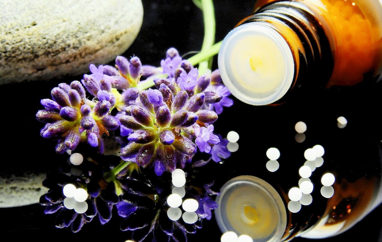 A close-up  displays homeopathic remedies, including a bottle of pills, white globules, and a sprig of lavender, alongside a smooth stone on a reflective black surface.
