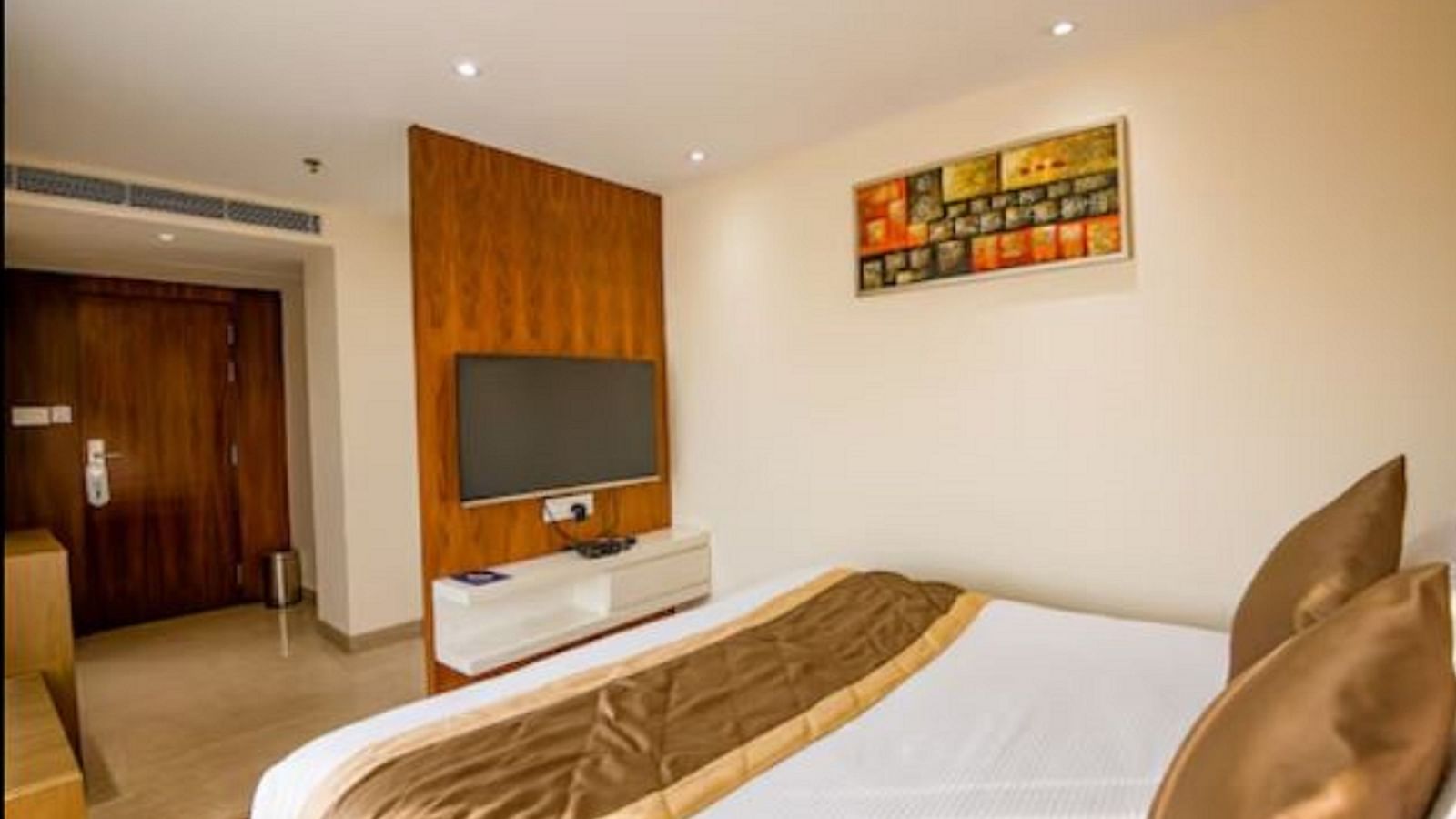 A neatly arranged standard hotel room with a large bed, a wooden headboard, a wall-mounted television, and a colourful painting above the bed at Yashshree The Imperial Inn, Siliguri.