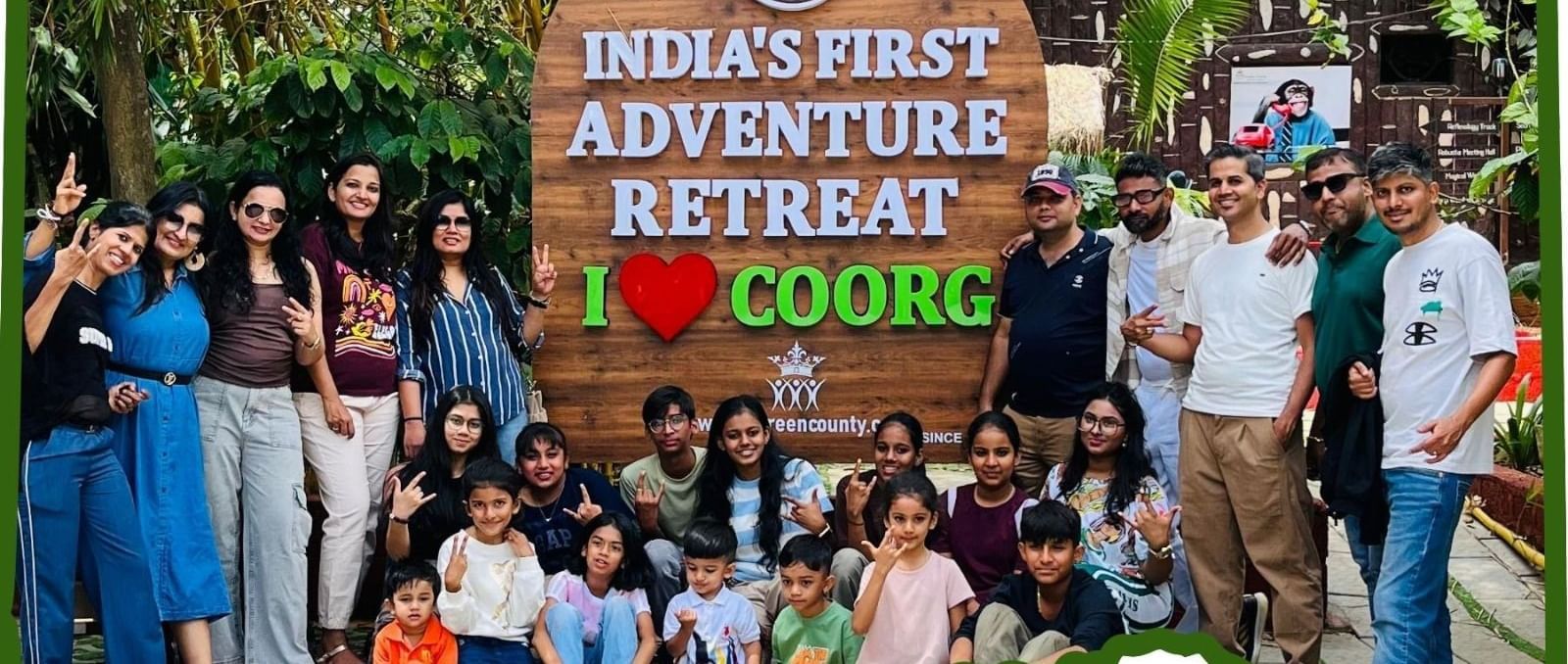 A group of people happily posing in front of a huge wooden board reading Indias First Adventure Retreat and I Love Coorg