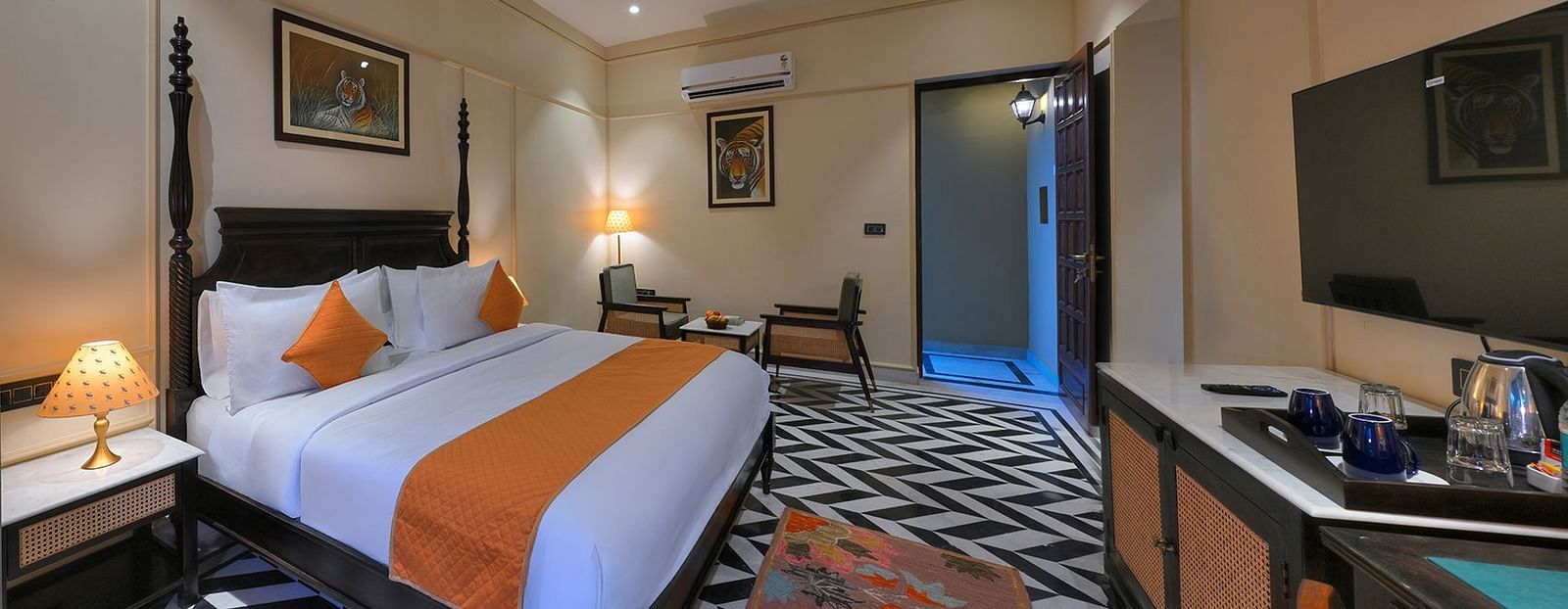 An interior view of Superior Cottage featuring a dark wood four-poster bed with white linens and orange accents, a flat-screen TV, a desk, and artwork depicting tigers on the walls - Singhvilas, Ranthambore