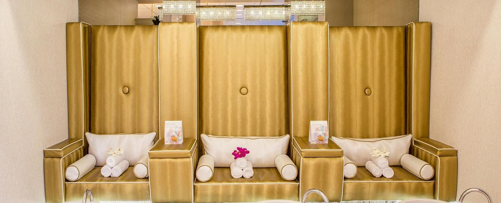 Spa pedicure setup with three plush gold chairs, each with towels and pillows, facing foot basins and a white countertop.