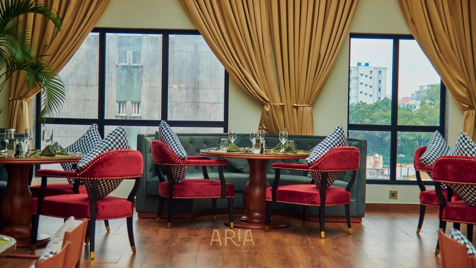 Elegant Aria restaurant interior with cushioned chairs, wooden tables, and large windows framed by curtains, allowing natural light to fill the space. City buildings and greenery are visible outside