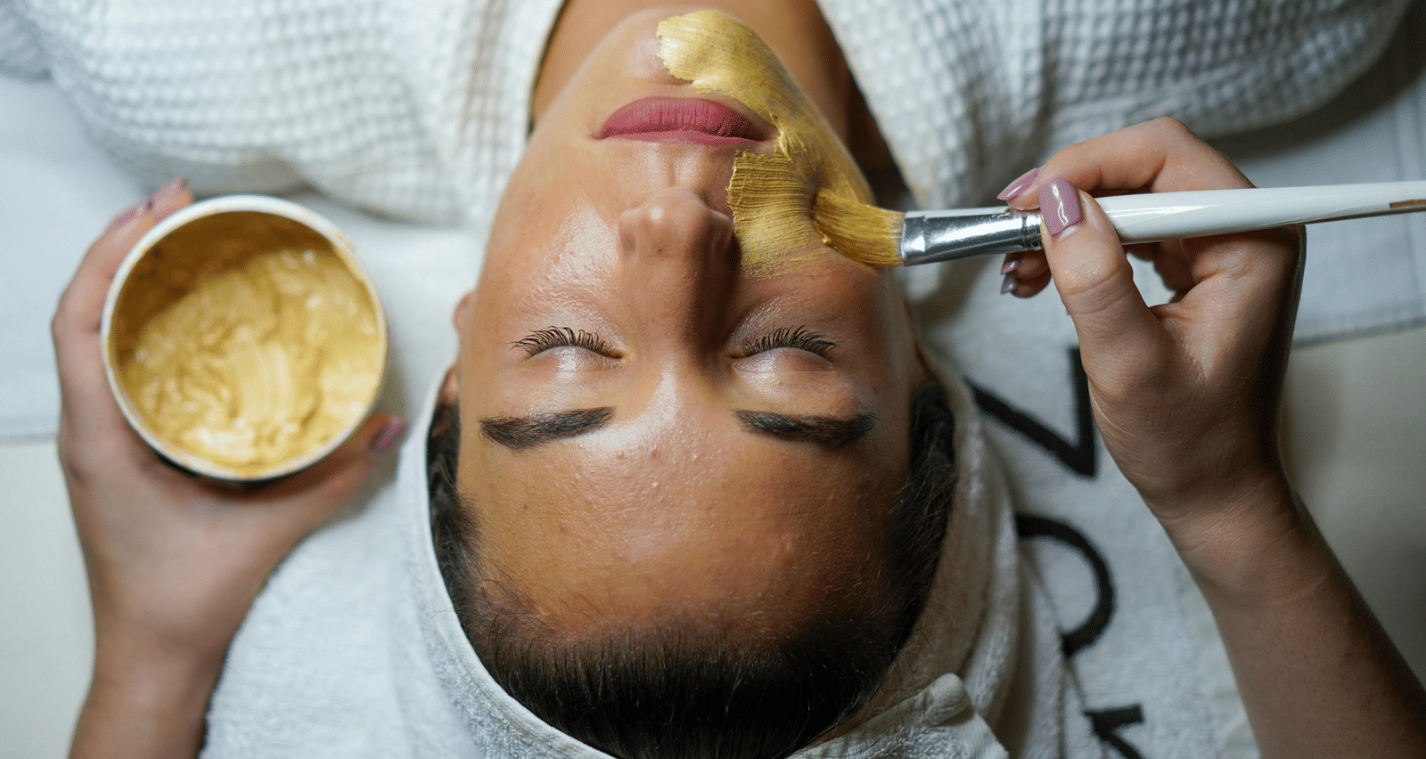 A person wearing a white robe is receiving a golden face mask application with a white brush.