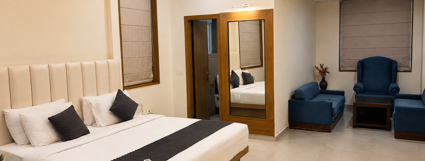 Suite at TGI Apple Inn, Ajmer Road, Jaipur featuring a bed with pillows, a seating area with armchairs and a table, blinds on the windows, and a doorway.