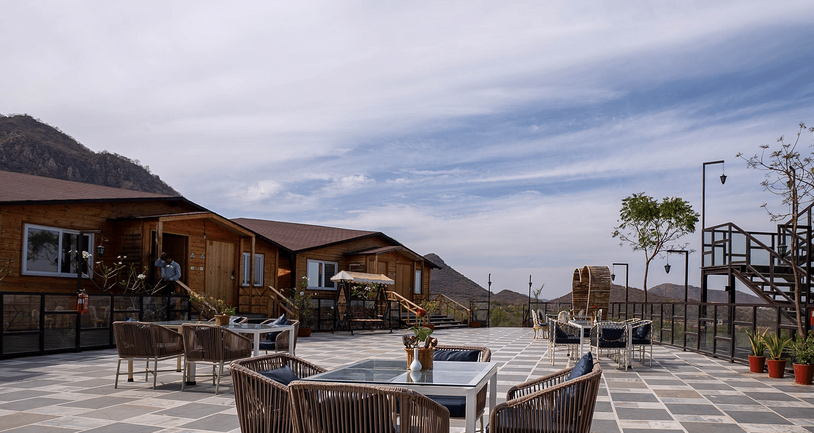 Outdoor patio with tables and chairs, wooden buildings in the background under a cloudy sky.- Bamboo Saa Resort & Spa, Udaipur