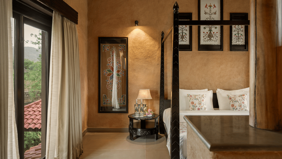 Cosy bedroom corner of Elegant Suite at AamaGhati Wildlife Resort featuring a dark wooden bed, earth-plastered walls decorated with framed floral motifs, and a glass door opening to a private balcony view.