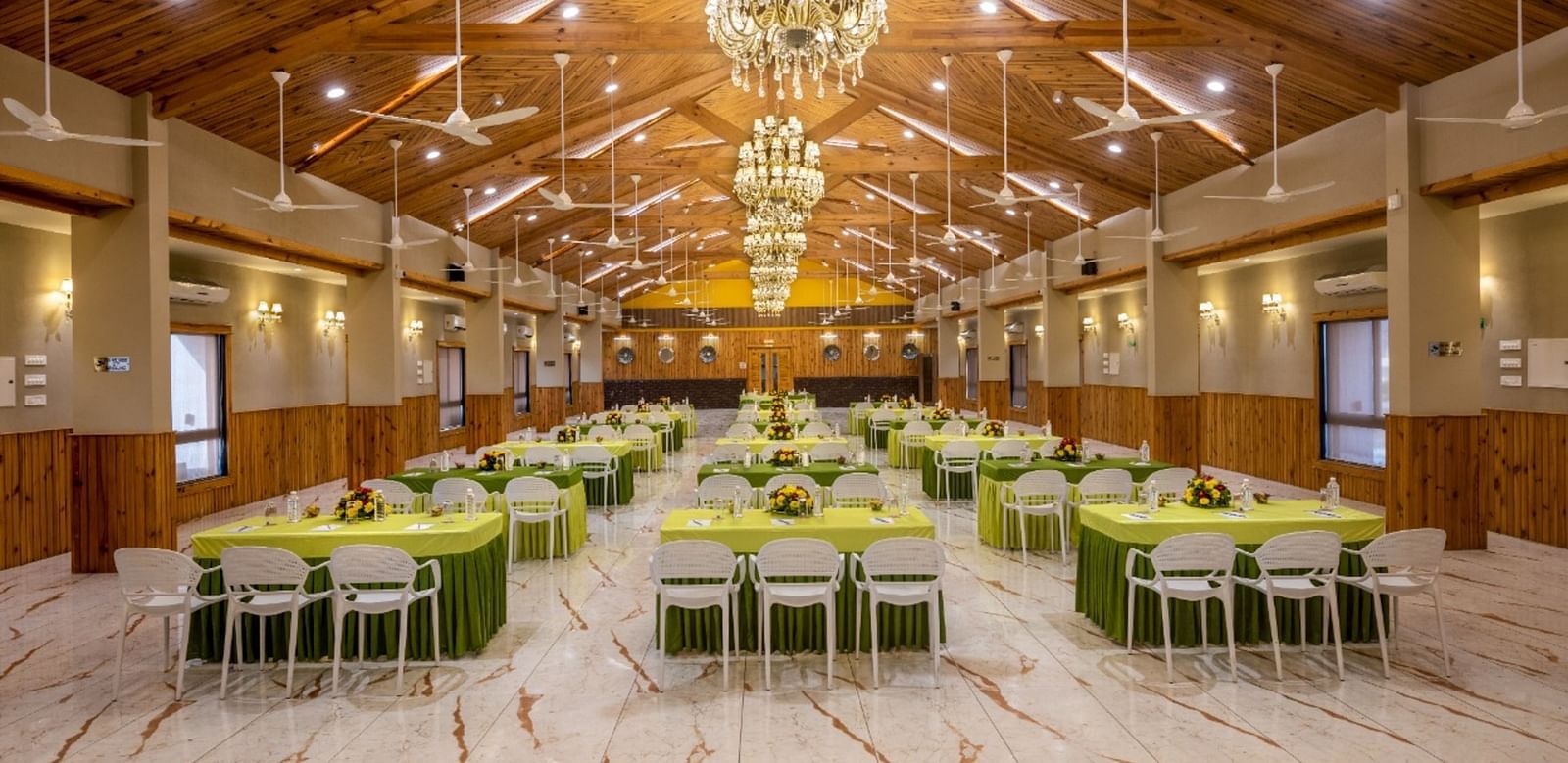 A well-lit banquet hall with multiple tables and chairs set for an event at Angel Lords Eco Inn Amreli.