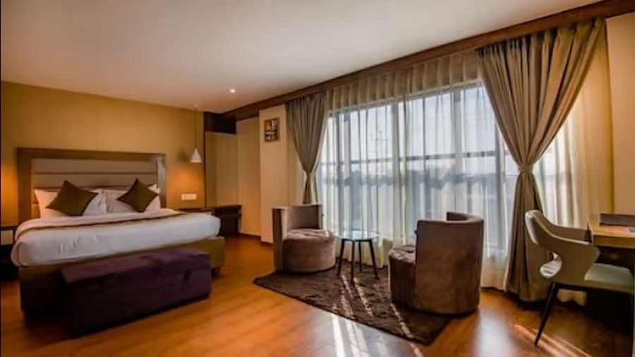 A premium hotel room with a large bed and windows that extend from the ceiling to the floor, with curtains pulled to the side, highlighting the two chairs and table at Yashshree The Imperial Inn, Siliguri.