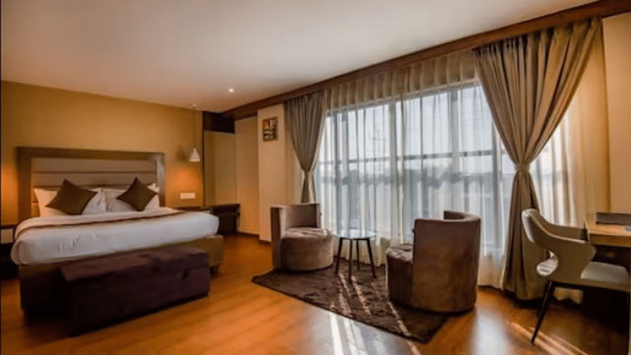 A premium hotel room with a large bed and windows that extend from the ceiling to the floor, with curtains pulled to the side, highlighting the 2 chairs and table at Yashshree The Imperial Inn, Siliguri