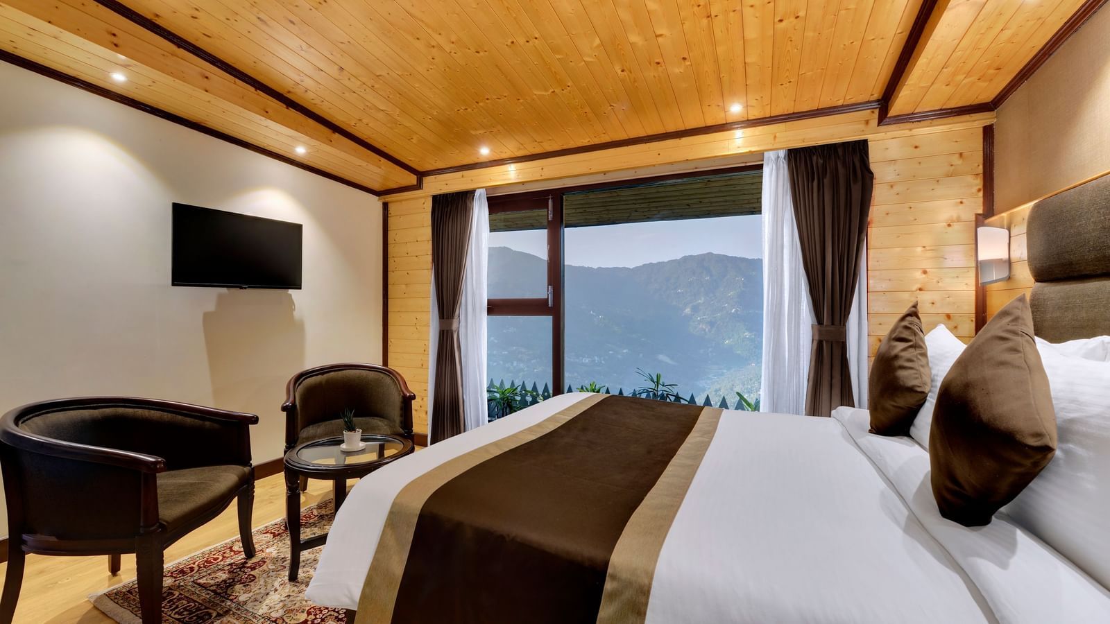 A premium triple room in Gangtok at the Sumitel Shangrila Deorali, featuring a queen-sized bed, two lounge chairs, a wall-mounted television opposite it, and a balcony.
