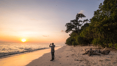 A person taking a picture of the rising sun at Kalapathar Beach while standing on the shoreline and trees next to him.