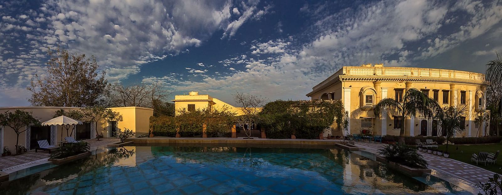  An outdoor scene featuring a pool reflecting the sky and buildings of Royal Heritage Haveli under a dramatic cloud formation.