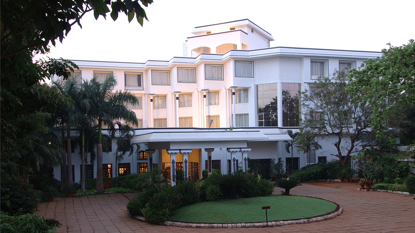 Facade view of Sangam Hotel, Thanjavur, with a pathway, a manicured garden and trees in view - Thanjavur Hotel Contact