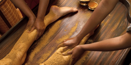 2 pairs of hands applying a substance on the legs of a person lying on a wooden surface