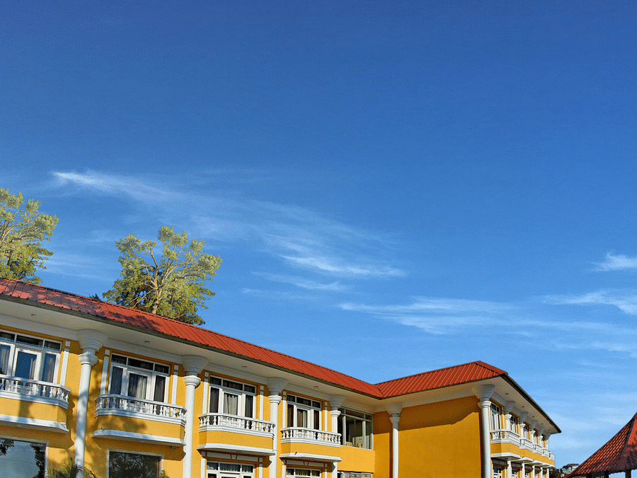 A corner exterior view of the  building with a roof and a balcony, surrounded by trees and a clear blue sky.