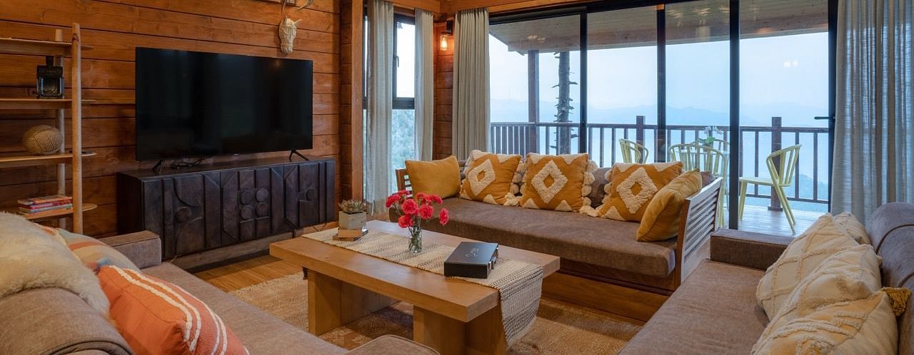 An image of a seating area with sofa sets, centre table, flat TV screen, chandelier lights at the ceiling, and a balcony attached to the room  -  Avalon Cottages, Kanatal 