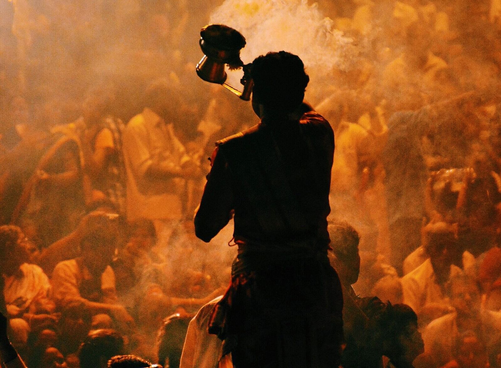 A close-up of a person performing Ganga Aarti, holding a lamp, surrounded by an engaged crowd. The scene captures the spiritual atmosphere