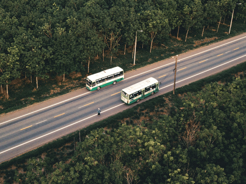 An aerial top-down view of 2 buses driving in opposite directions on a road cutting through a dense forest.