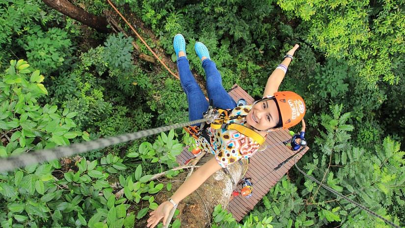 A person wearing a helmet and harness joyfully glides through the dense, bright green canopy of a forest while ziplining, with sunlight filtering through the leaves