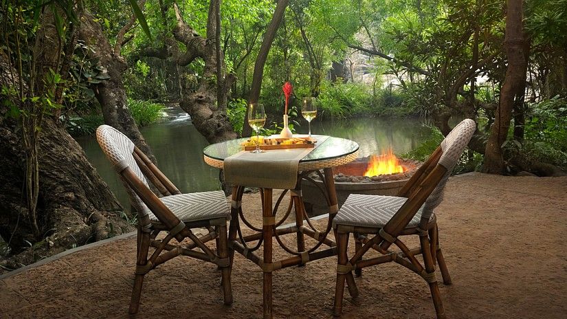 Riverside dining setup with lit candles and comfortable chairs, surrounded by trees.