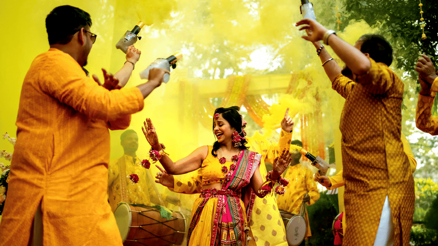 A couple in yellow outfits celebrating a Haldi ceremony at Vedic Village Spa Resort with yellow color powder in the air surrounded by guests in a joyful outdoor setting.
