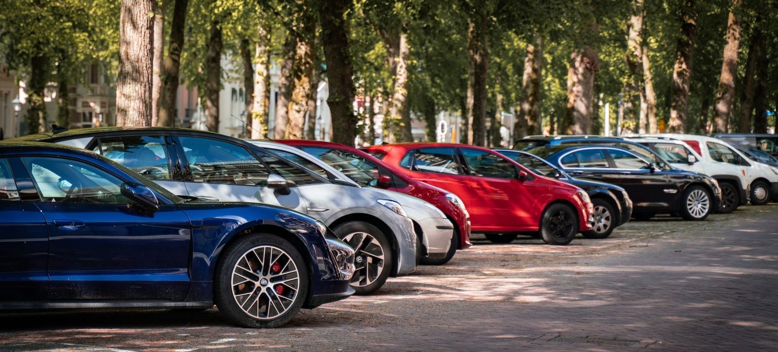 A row of sleek, modern cars, including a blue, silver, and red model, are parked along a tree-lined street, casting shadows on the pavement.
