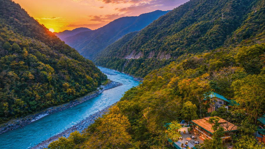 Aerial view of the Neemran's Glasshouse on The Ganges, Rishikesh, surrounded by lush greenery by the river banks, along with a mesmerising sunset in the background.