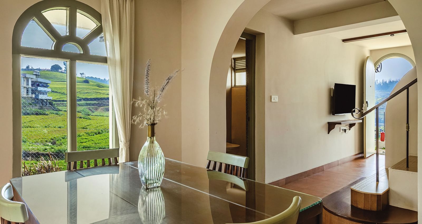 dining space at Mango Hill Shola, Ooty, featuring elegant furniture and a large arched window that frames a  hillside & building view.
