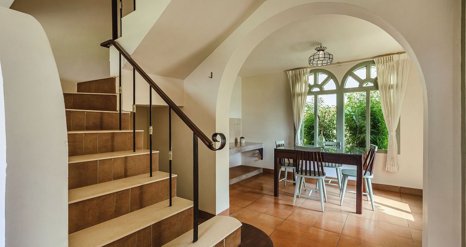 Interior view at Mango Hill Shola, Ooty, showing a wooden staircase, a dining table, and arched windows in the background.