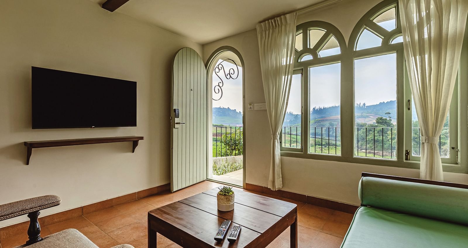 Inviting living area at Mango Hill Shola, Ooty, featuring a mounted television, cushioned seating, and large arched windows letting in ample natural light.