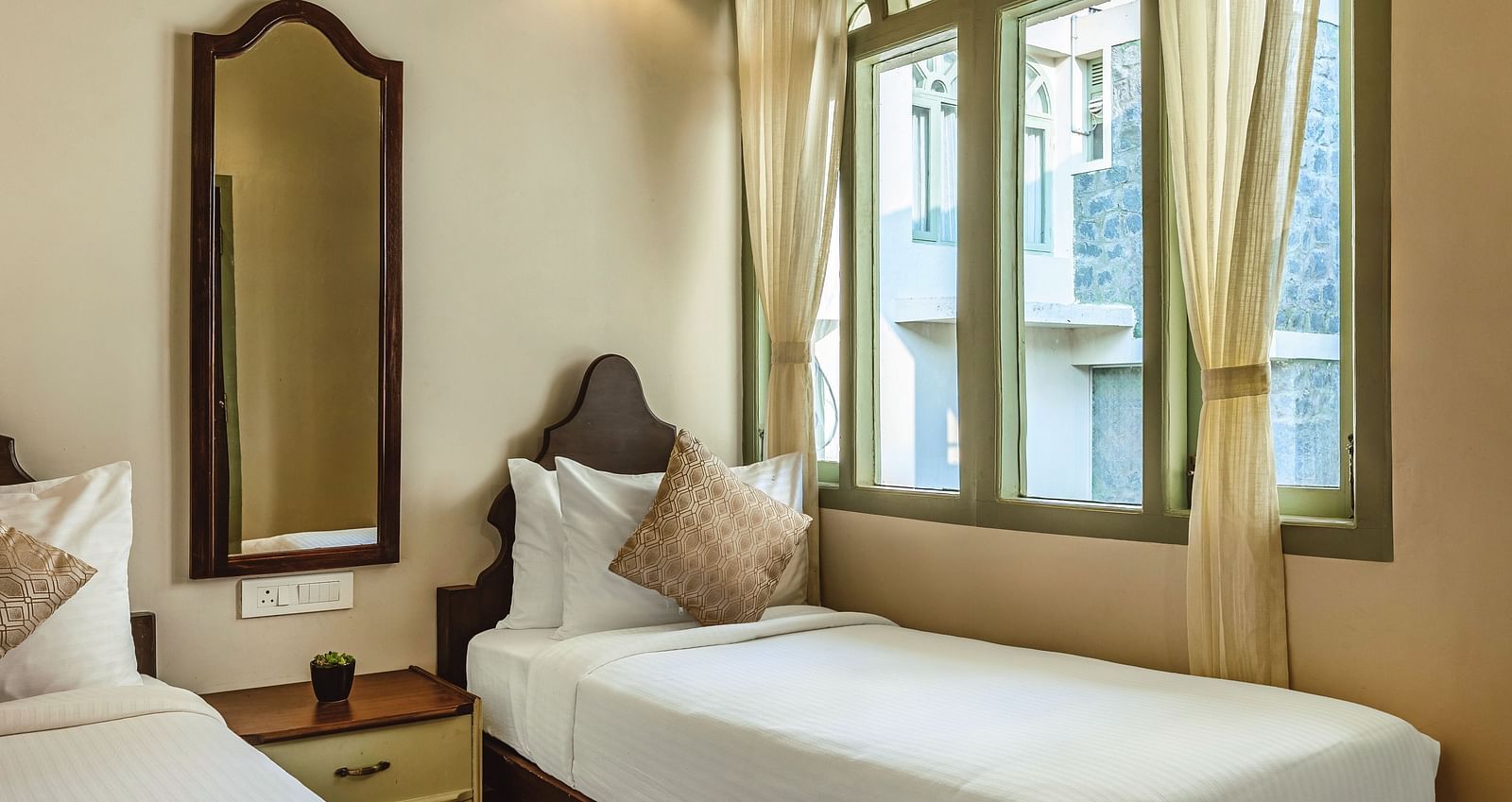 Elegant bedroom at Mango Hill Shola, Ooty, featuring a double bed with crisp white linens, wooden bedside tables, and a tall arched window offering views of the lush landscape.