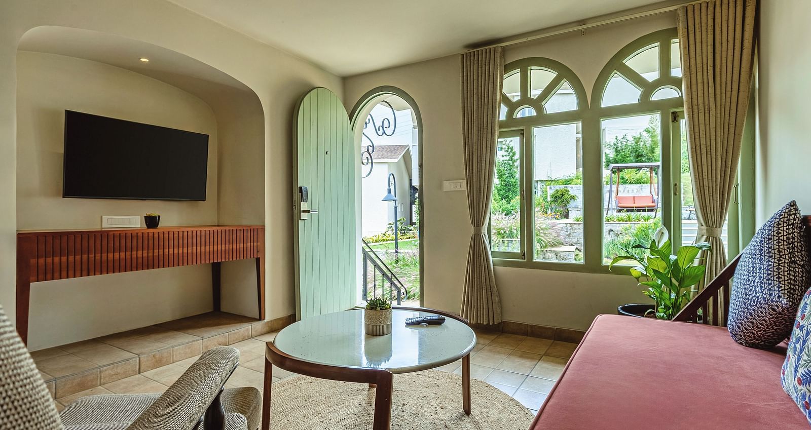 Spacious living room with a red sofa, TV, and arched windows overlooking the garden at Mango Hill Shola, Ooty.