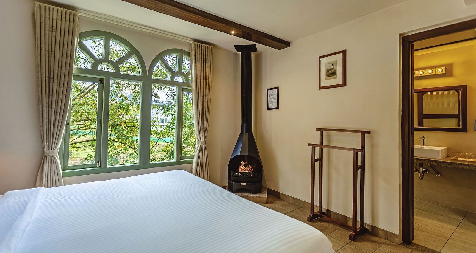  A corner view of a warm and elegant bedroom at Mango Hill Shola, Ooty with a central fireplace and peaceful outdoor scenery visible through the windows.