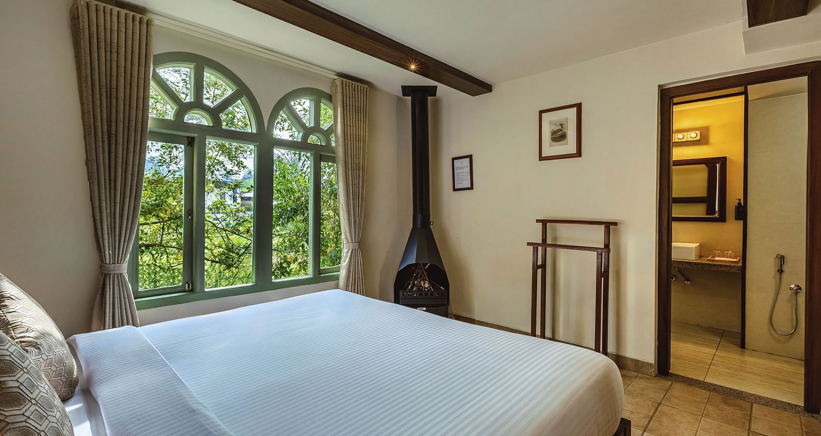 Well-lit bedroom with a queen bed, classic furniture, and garden views through a large arched window at Mango Hill Shola, Ooty.