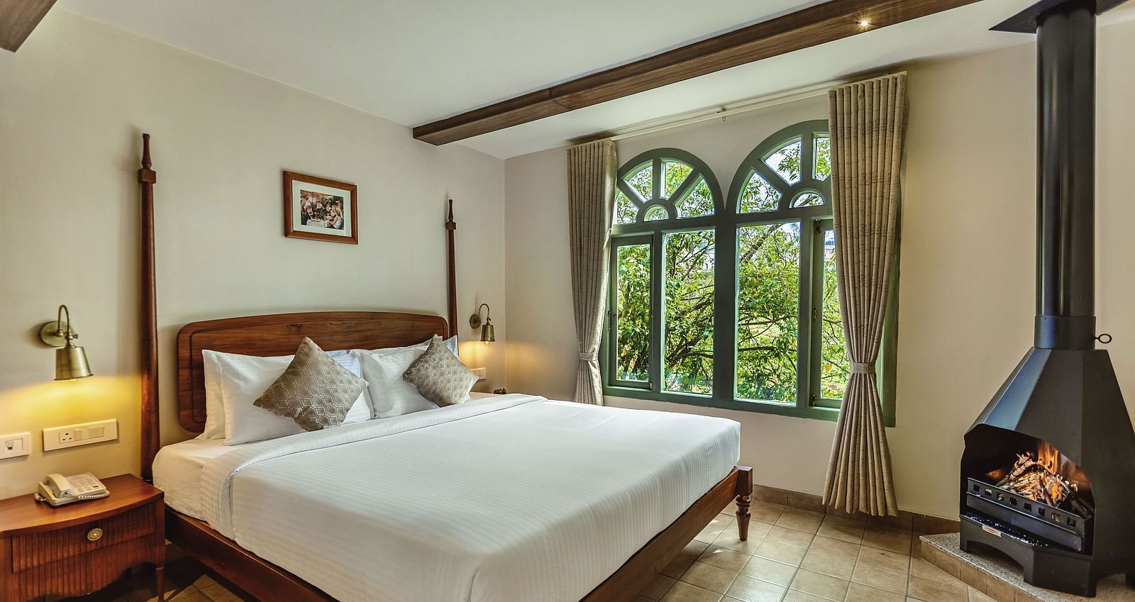 Sunlit bedroom at Mango Hill Shola, Ooty with a wooden bed, side tables, and large arched windows offering lush green views.