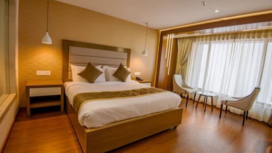 The image displays a deluxe hotel room with a large bed with a wooden headboard and a bedside table. It has a huge window to the side, along with two chairs and a mini table between them at Yashshree Imperial, Siliguri.