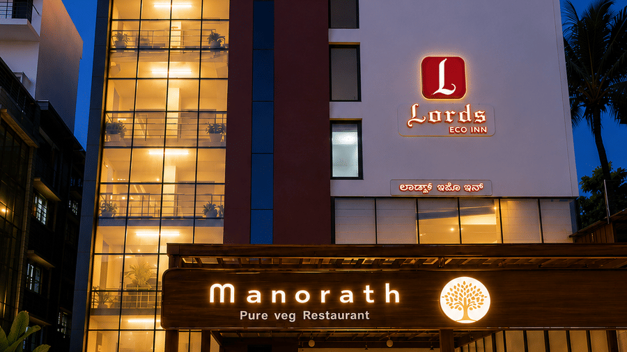 Night view of an illuminated multi-story hotel building featuring a glass-paneled stairwell, warm exterior lighting, and the ground-level entrance to Manorath Fine Dining Restaurant - Lords Eco Inn Belagavi