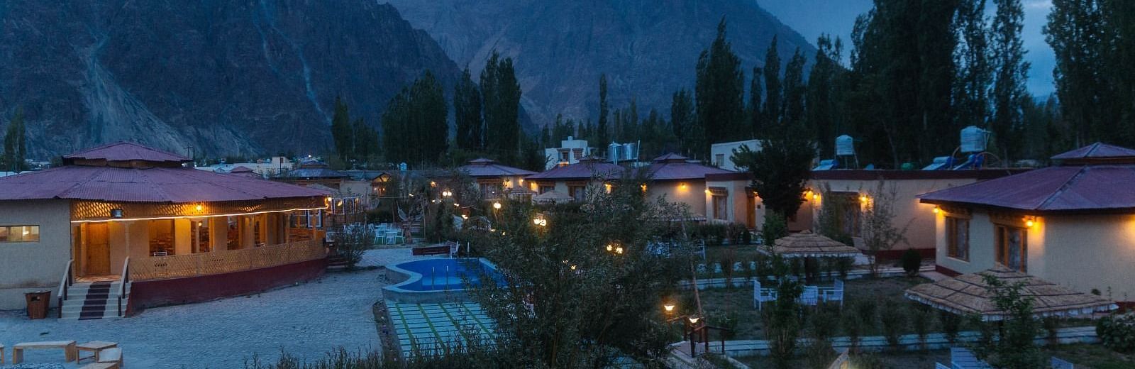 Aerial view of White Sand Resort, Ladakh at dusk, showing cabins, trees, and mountains under a cloudy sky.