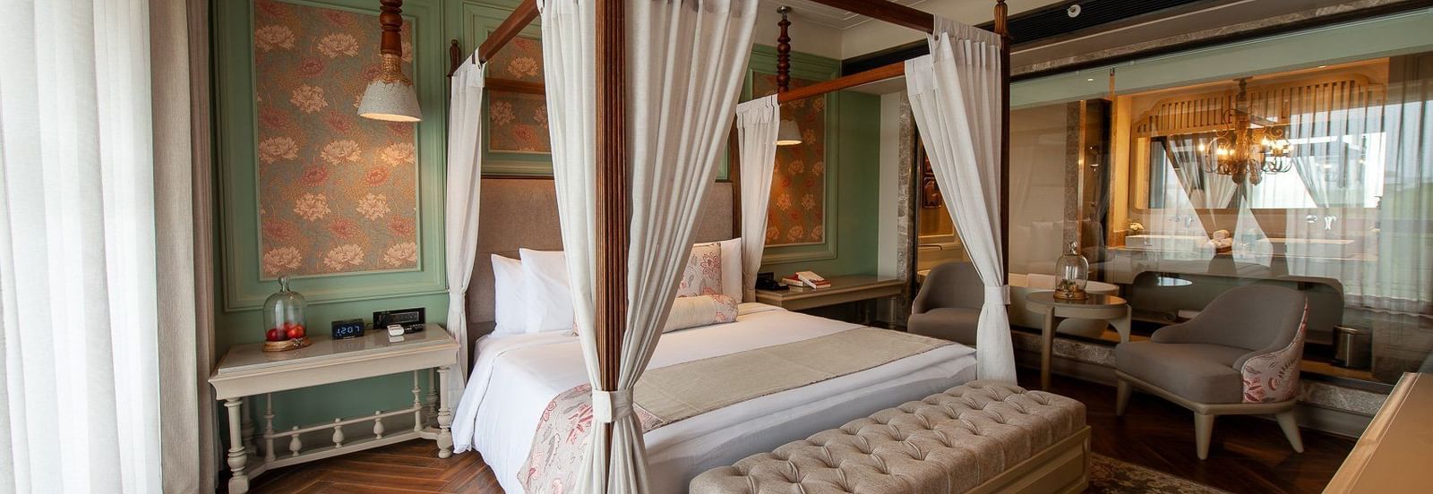 A wide view of the elegant suite at The Evren, Vagator, showcasing a four-poster bed, a stylish rug on a herringbone floor, and a seating area with two armchairs near a window. The room has a view into the connected bathroom.