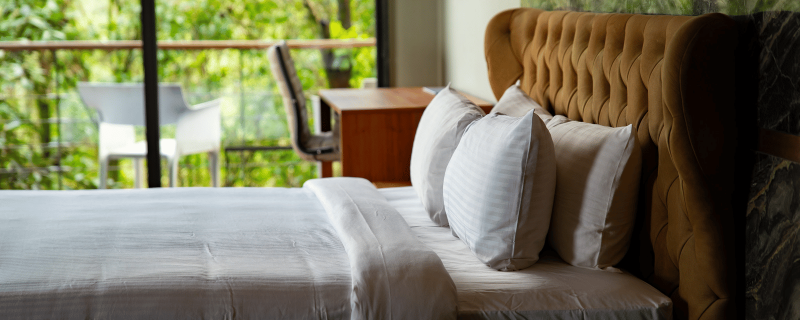 A close-up of a large bed with a high, tufted headboard at Stanley Revelation, with a forest visible outside the window.